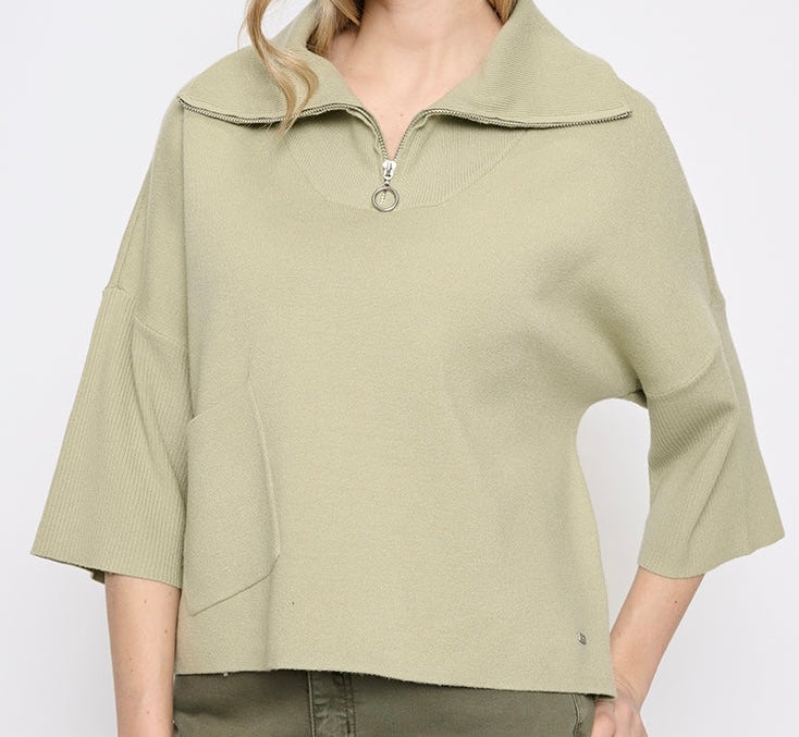 A woman wearing a light sage green ribbed knit top with a half-zip collar and elbow-length sleeves, shown against a plain light gray background. -OLIVIA'S