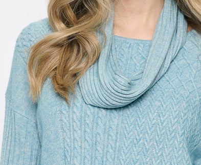 A close-up of a woman wearing a light blue cable-knit cowl neck sweater, showing her blonde hair and the textured knit pattern. - OLIVIA'S