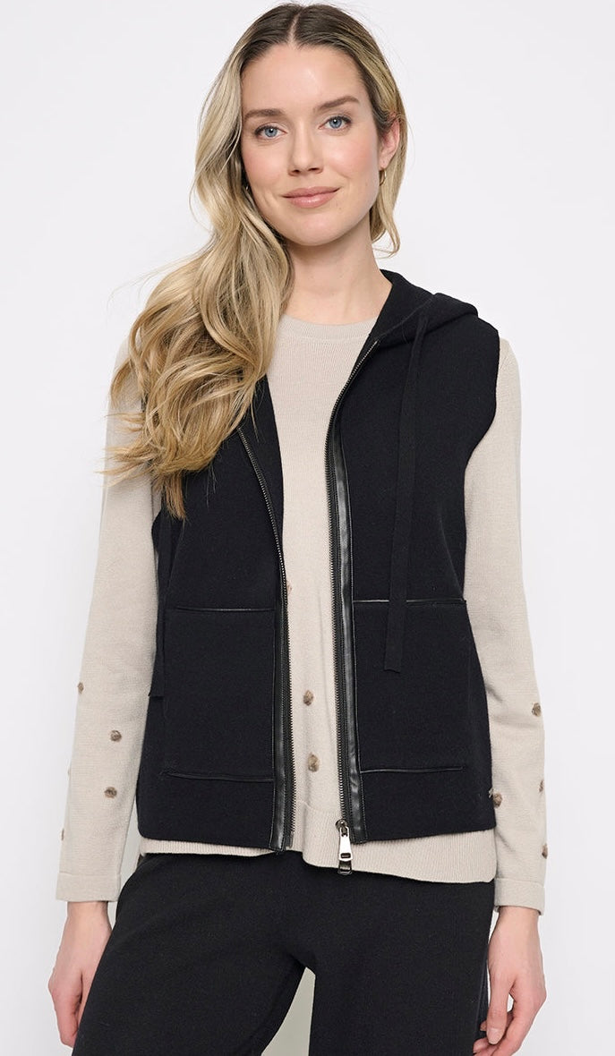 A woman modeling a black sleeveless zip-front vest with a hood over a light beige long-sleeve top and black pants, photographed against a plain light gray studio background. - OLIVIA'S