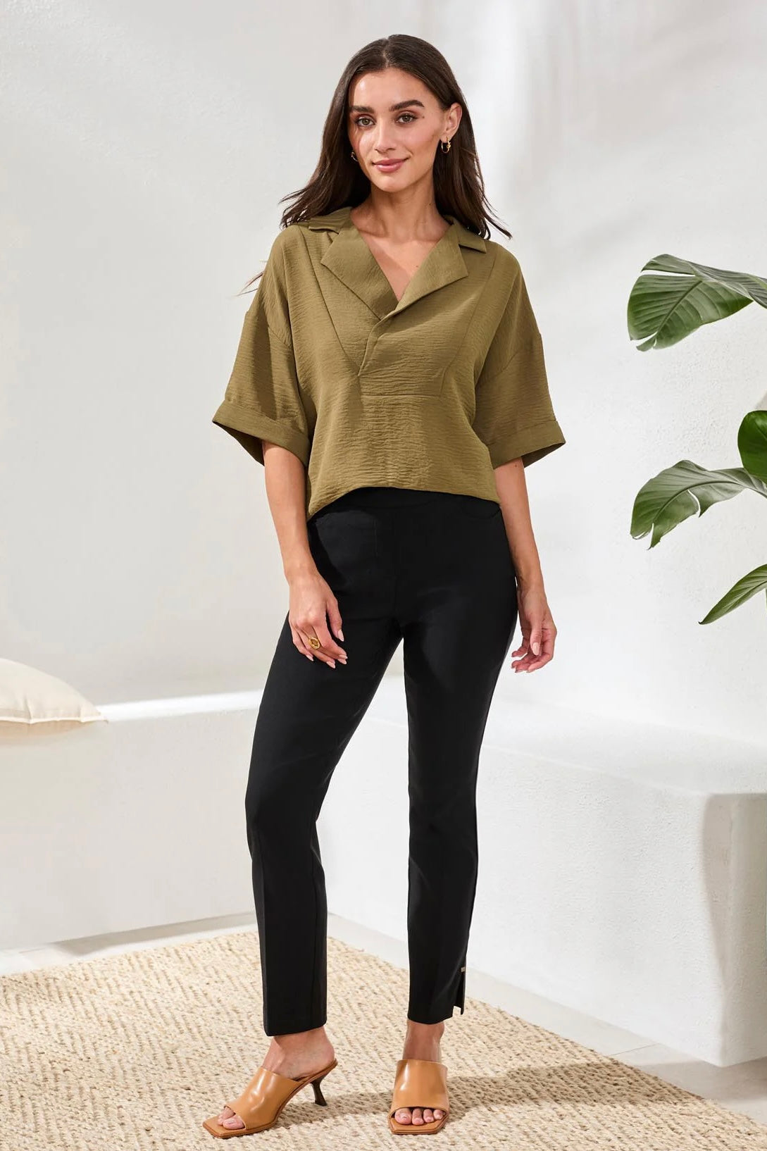 Woman wearing a green top and black pants standing in a minimalistic room with a plant.