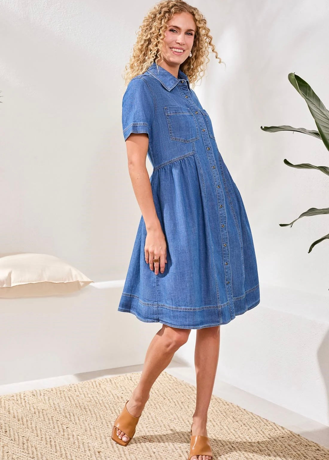 Woman wearing a blue denim dress standing in a room with a white wall and plant.