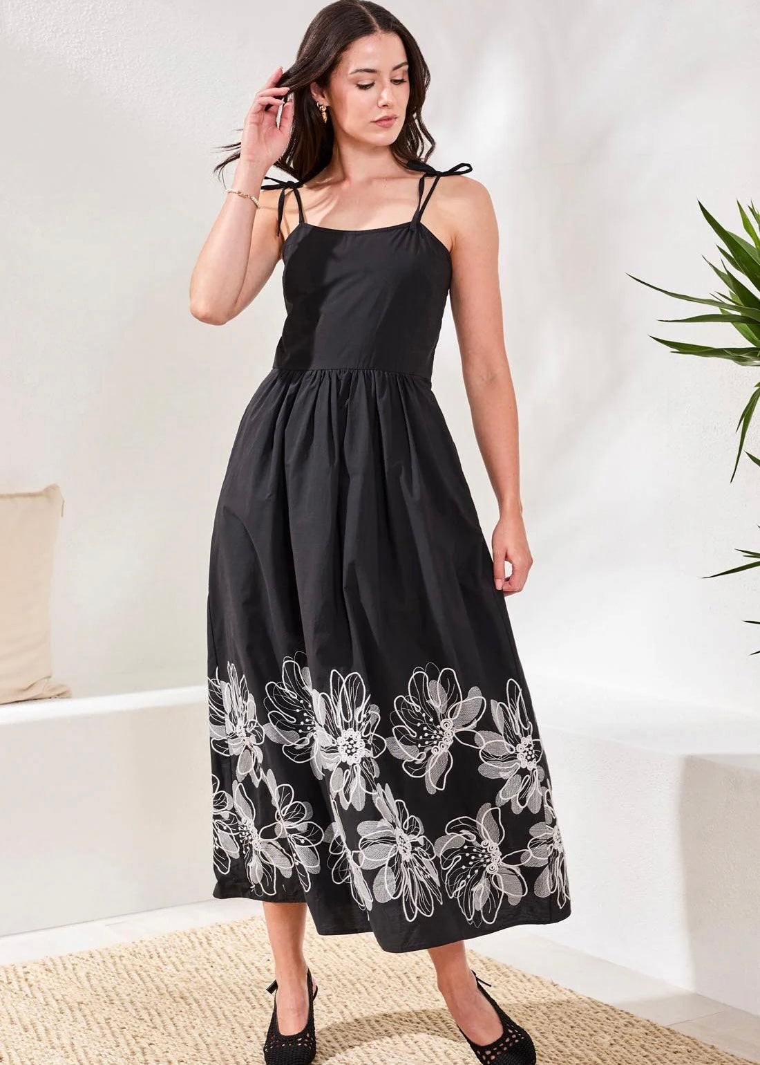 Woman wearing a black dress with white floral embroidery in a minimal indoor setting.