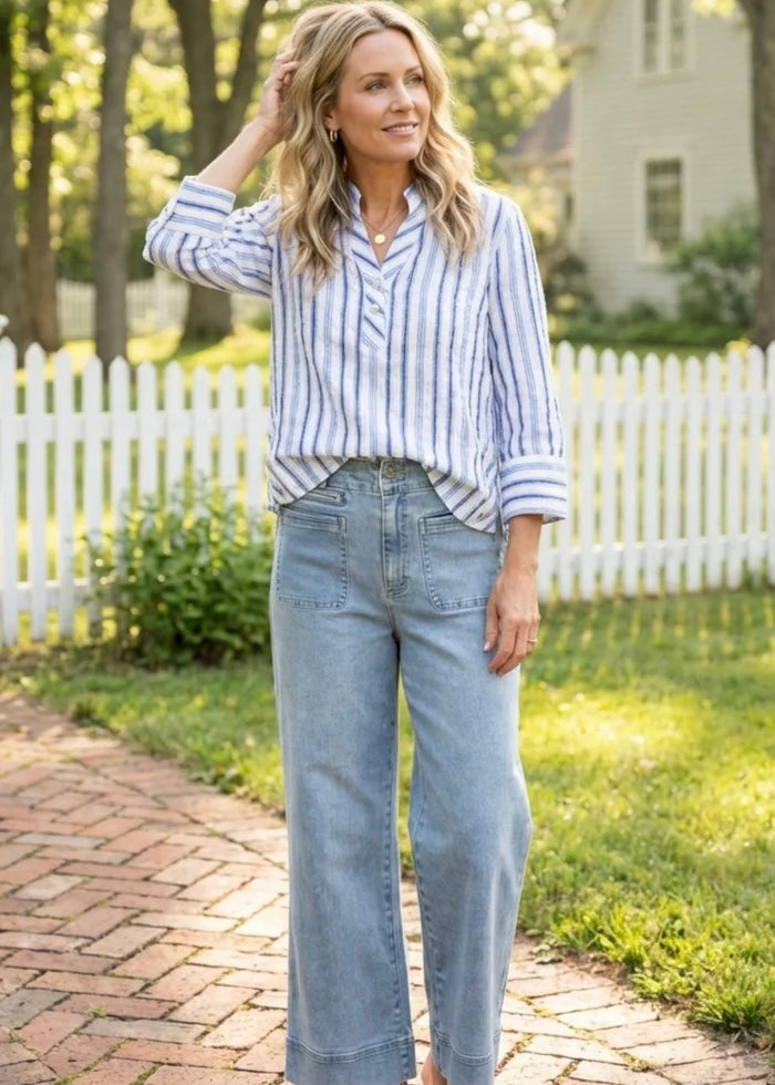 Woman wearing a striped shirt and jeans standing on a brick path with a white picket fence and greenery in the background.