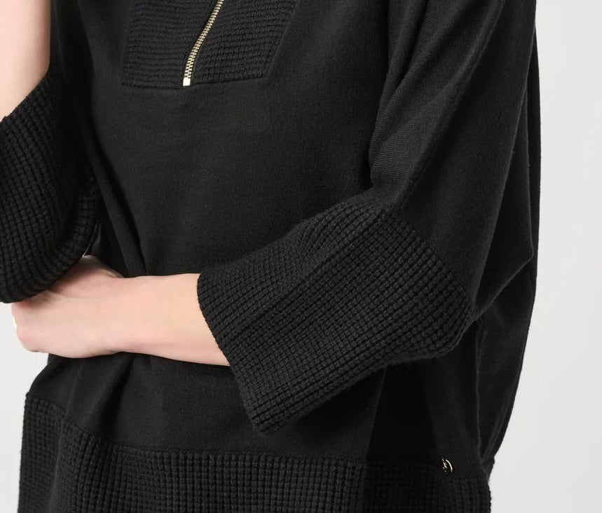 Close-up of a black knit top with textured ribbed cuffs and a small gold zipper detail on the chest, worn by a model against a light gray background.