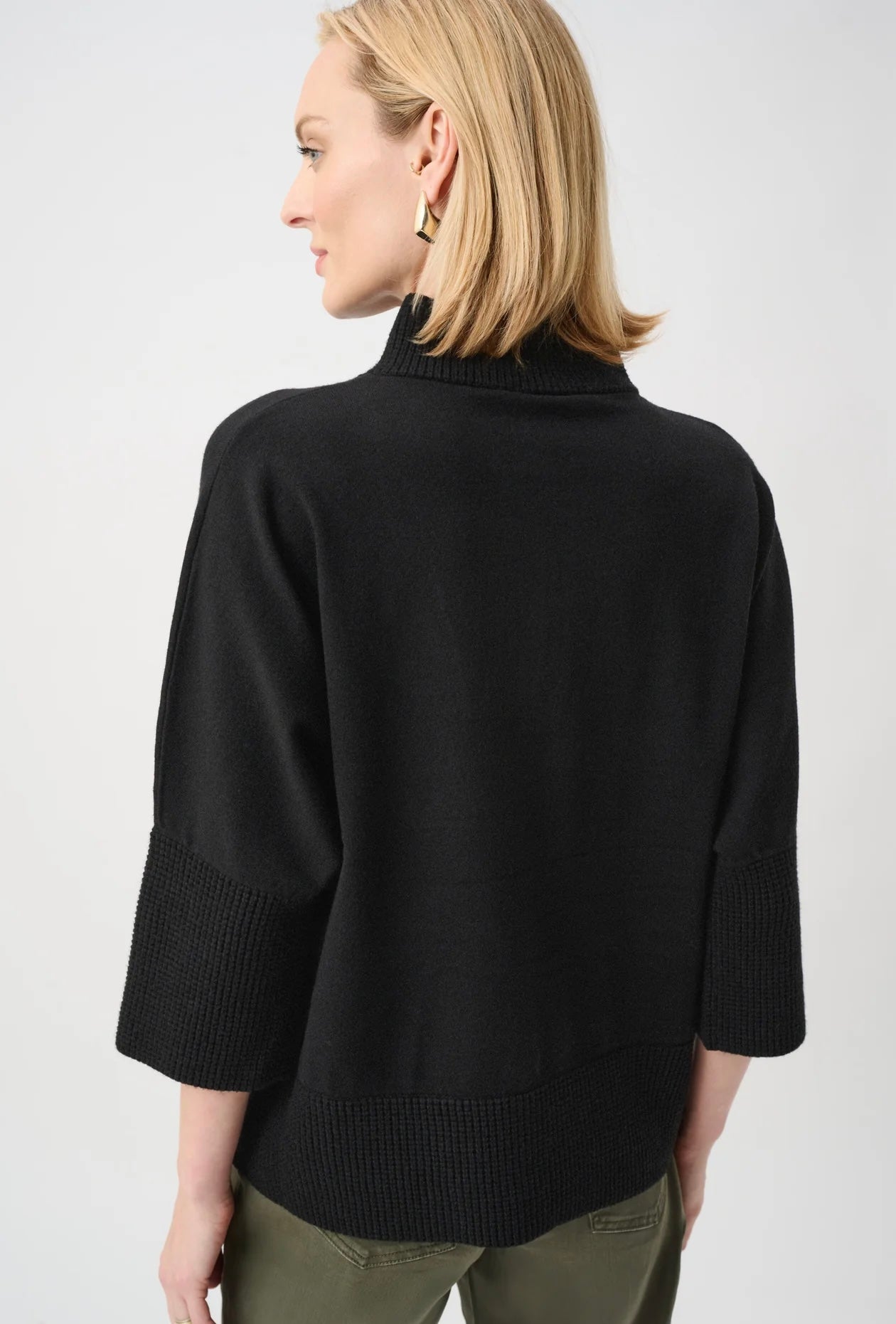 A woman shown from the back wearing a black knit mock-neck sweater with ribbed three-quarter sleeves and hem, paired with olive green pants, against a light gray studio background.