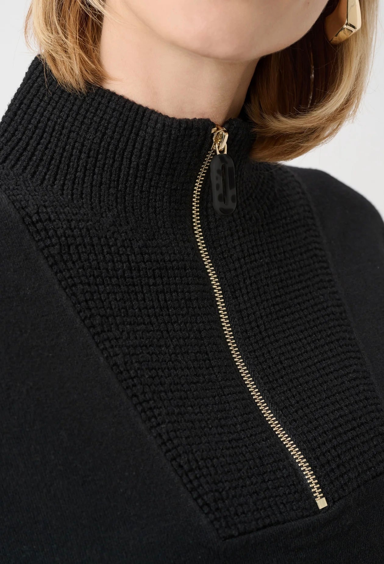 Close-up of a black ribbed knit sweater with a high mock neck and a prominent gold zipper running down the center front, worn by a model.
