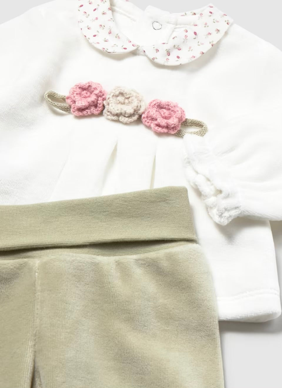 A baby girl's two-piece outfit featuring a white long-sleeve top with a floral-patterned Peter Pan collar and a decorative pink and beige flower hair clip, paired with light olive green corduroy pants.