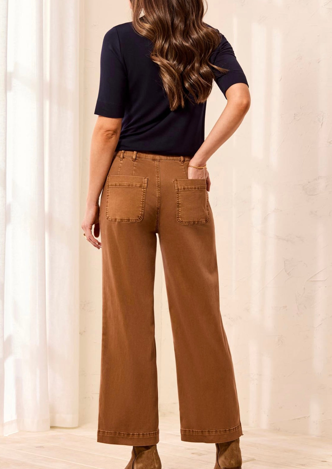 A woman shown from the back wearing brown wide-leg pants, a black short-sleeve top, and brown boots in a brightly lit room with white curtains.