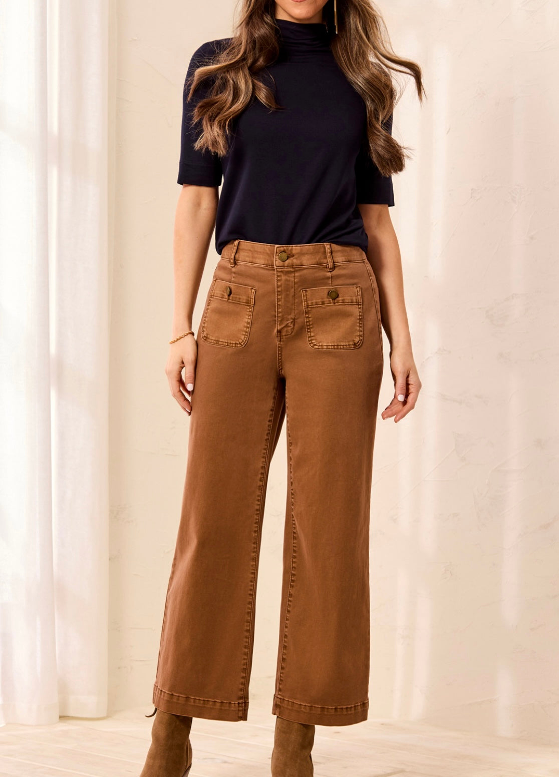 A woman wearing brown wide-leg cropped jeans with front patch pockets, styled with a black short-sleeve mock neck top and brown ankle boots, standing in a brightly lit indoor setting.