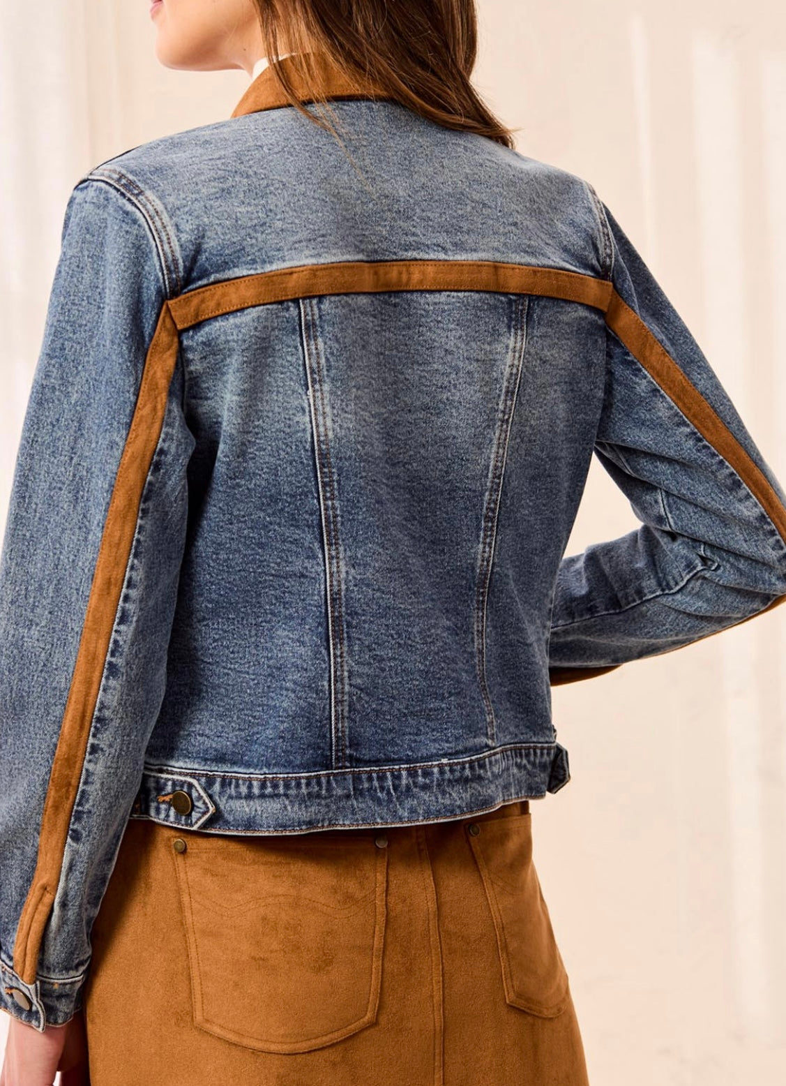 A woman shown from the back wearing a cropped blue denim jacket with contrasting brown suede-like shoulder and sleeve panels, paired with brown pants.