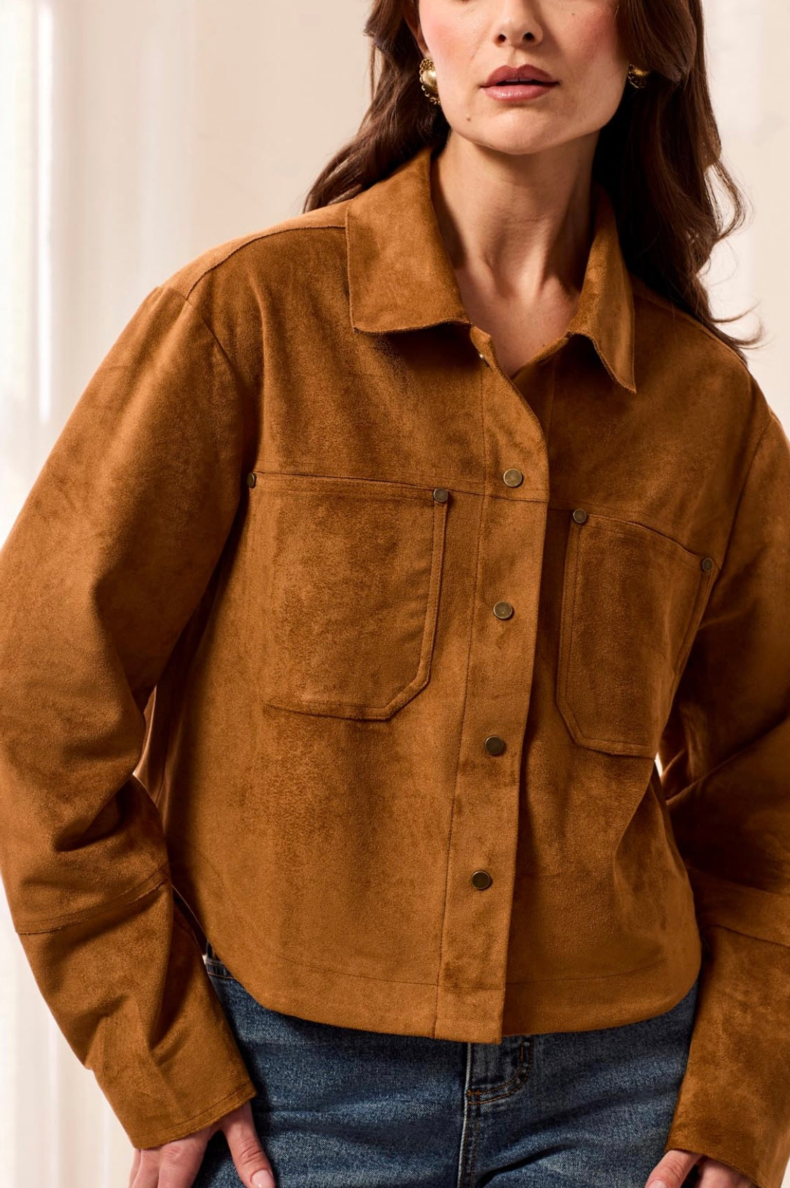 A woman wearing a cropped, long-sleeve suede or faux-suede button-up shirt jacket in a warm camel brown color, paired with blue jeans.