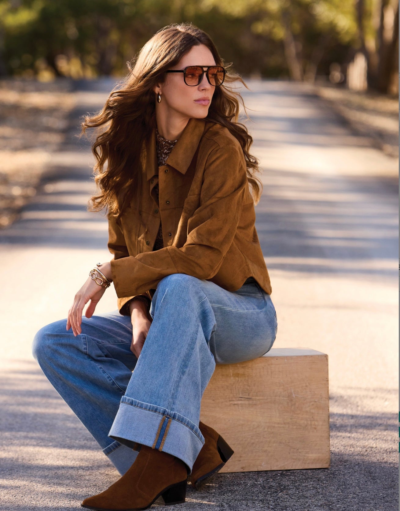 A woman sits on a wooden block outdoors on a sunlit road, wearing a brown suede jacket, light blue jeans, brown ankle boots, and sunglasses.