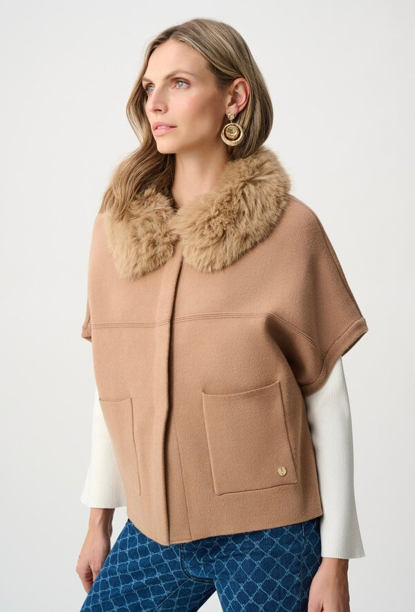 A woman wearing a camel-colored cape jacket with a large tan faux-fur collar, layered over a white long-sleeve top and blue patterned pants, photographed against a light gray studio background.