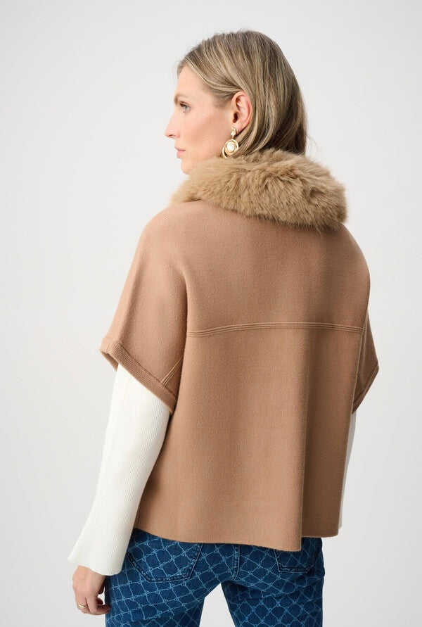 A woman shown from the back wearing a camel-colored knit cape with a fluffy tan faux-fur collar, layered over a white long-sleeve top and blue patterned pants, against a light gray studio background.