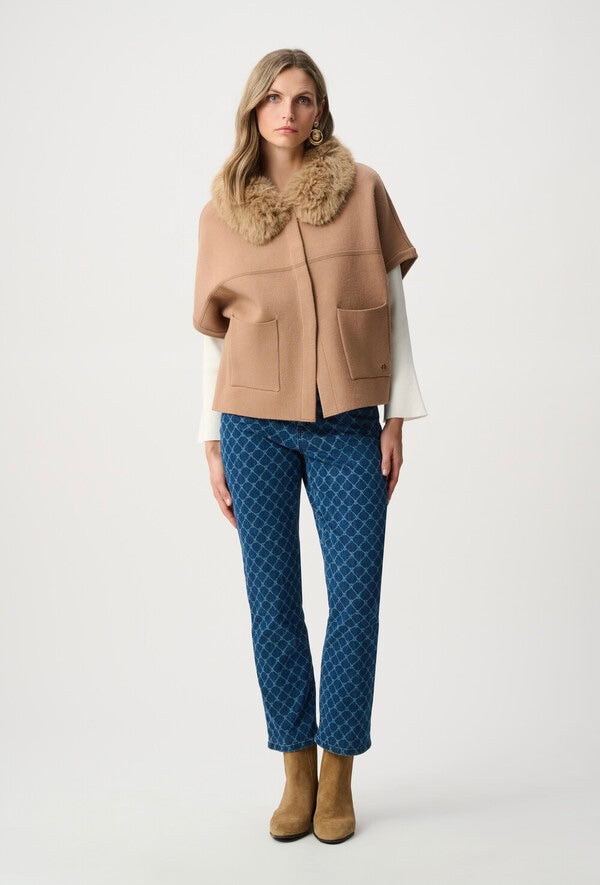 A model stands against a light gray studio background wearing a camel-colored cape jacket with a fluffy beige fur collar, a white long-sleeve shirt, blue patterned trousers, and tan ankle boots.