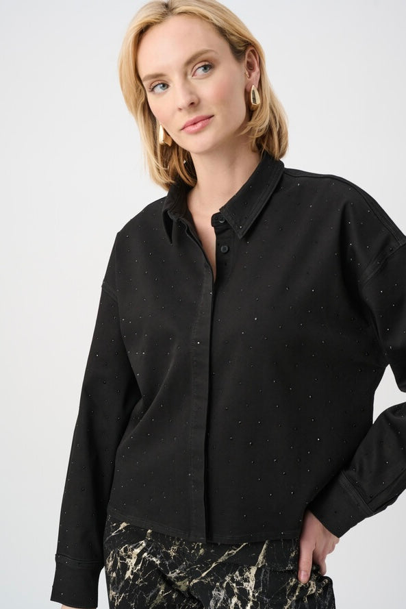 A woman wearing a black long-sleeve button-up shirt with subtle silver speckles, paired with black and beige patterned trousers, photographed against a light gray studio background.