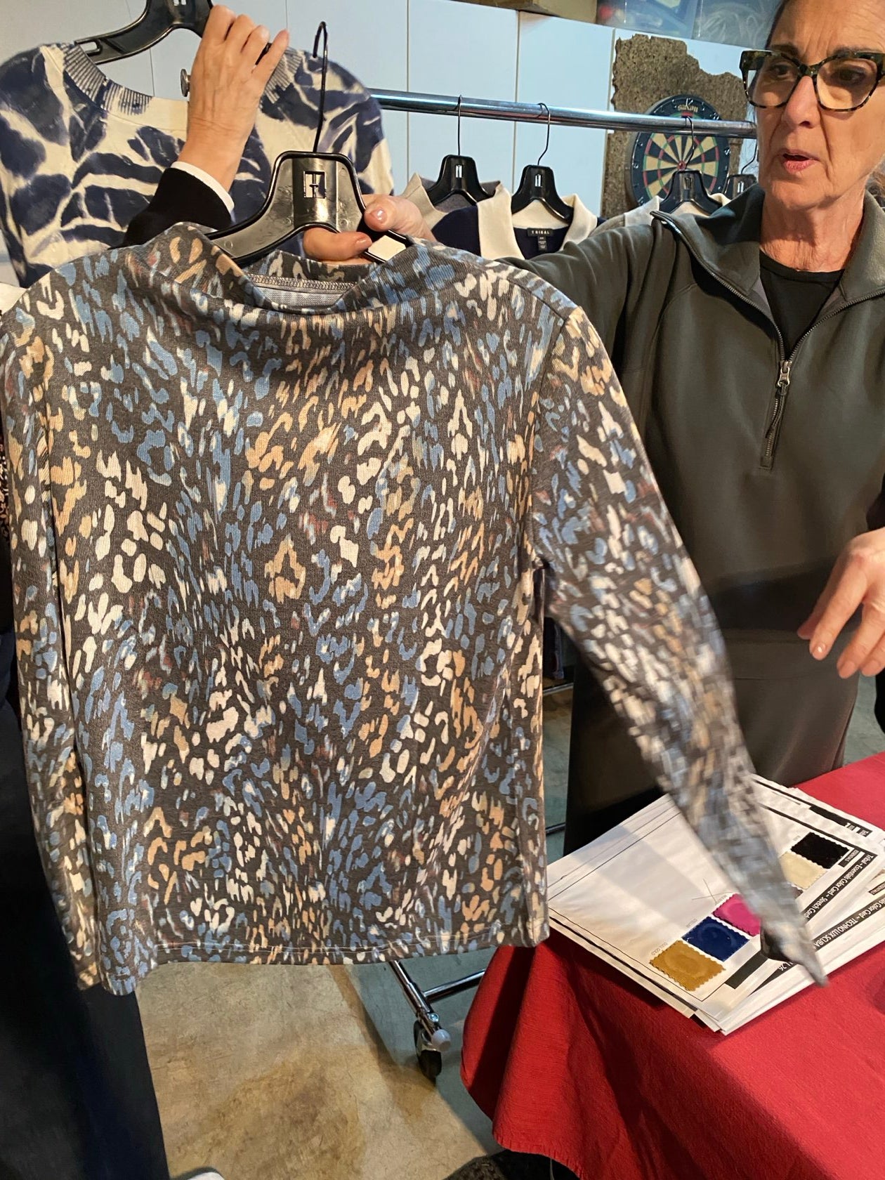 A person holding up a long-sleeve fleece pullover with an abstract leopard-like pattern in beige, brown, and blue-gray tones. The photo is taken in a retail or studio setting with a red tablecloth and other garments visible in the background.
