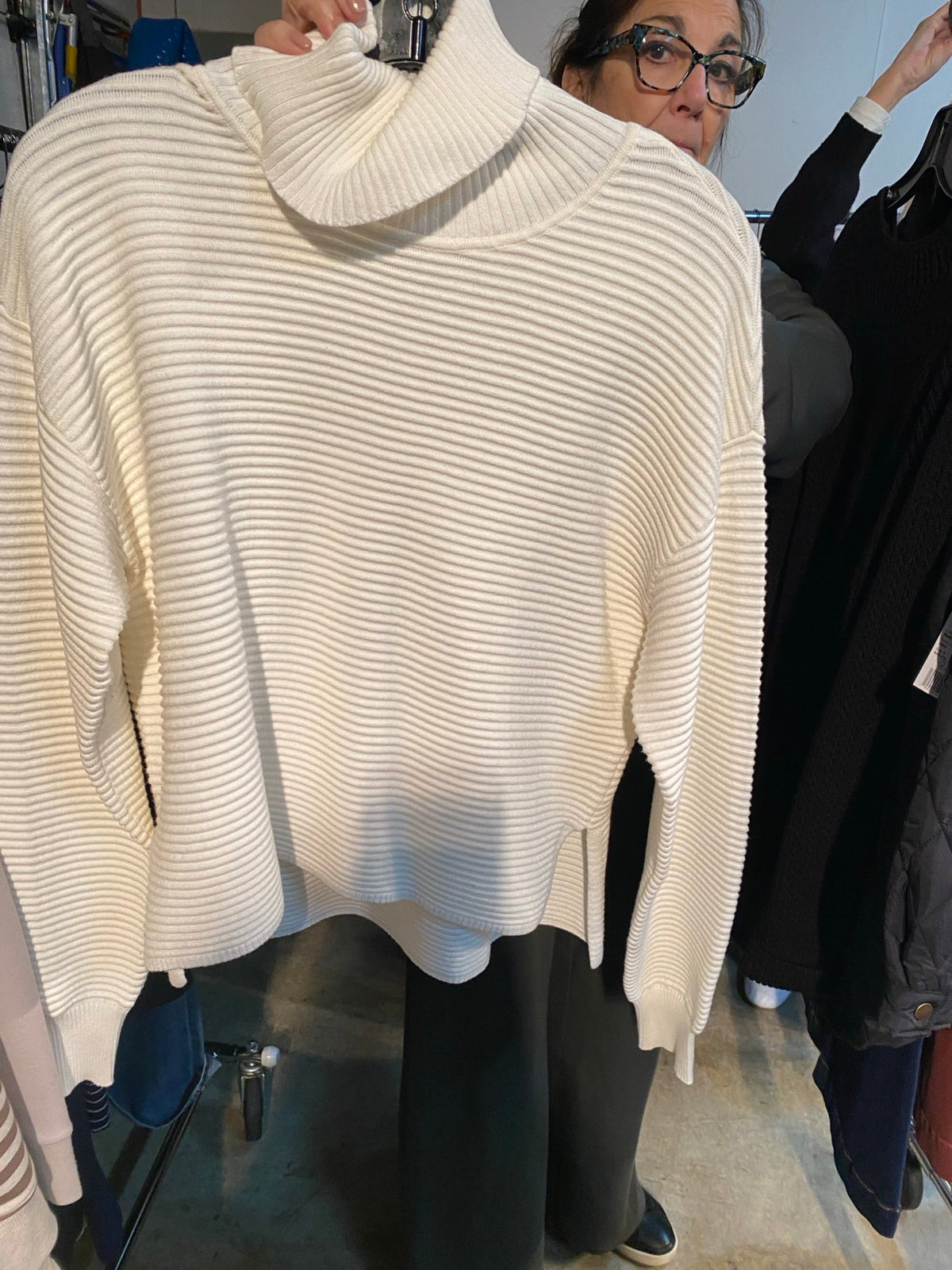 A cream-colored ribbed knit sweater with a high turtleneck collar, shown on a person in a clothing store setting. - OLIVIA'S