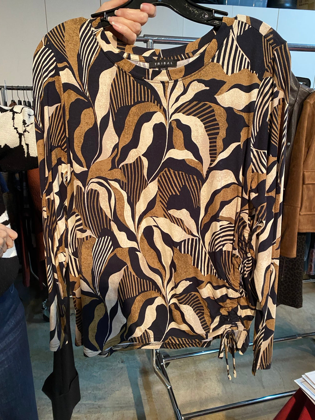 A long-sleeve knit top with a bold abstract botanical pattern in black, tan, and cream, held up on a hanger by a person in a clothing store.