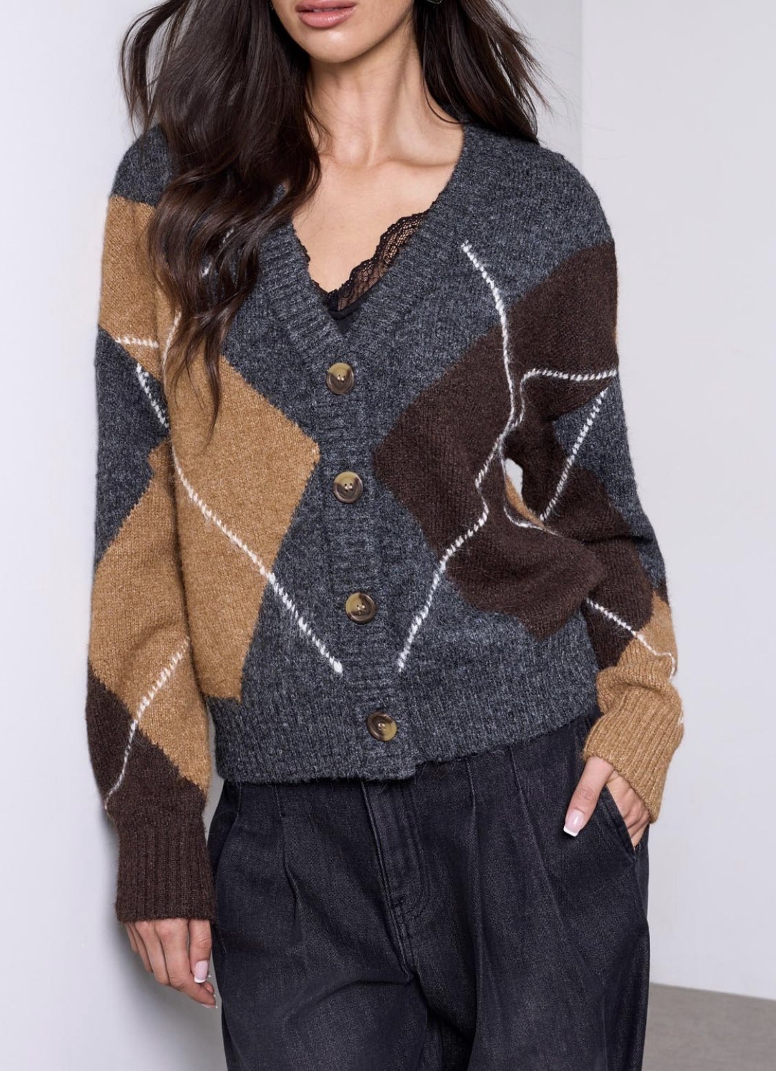 A woman wearing a chunky knit cardigan with a geometric argyle pattern in dark gray, brown, and tan, featuring brown buttons and a lace V-neckline.