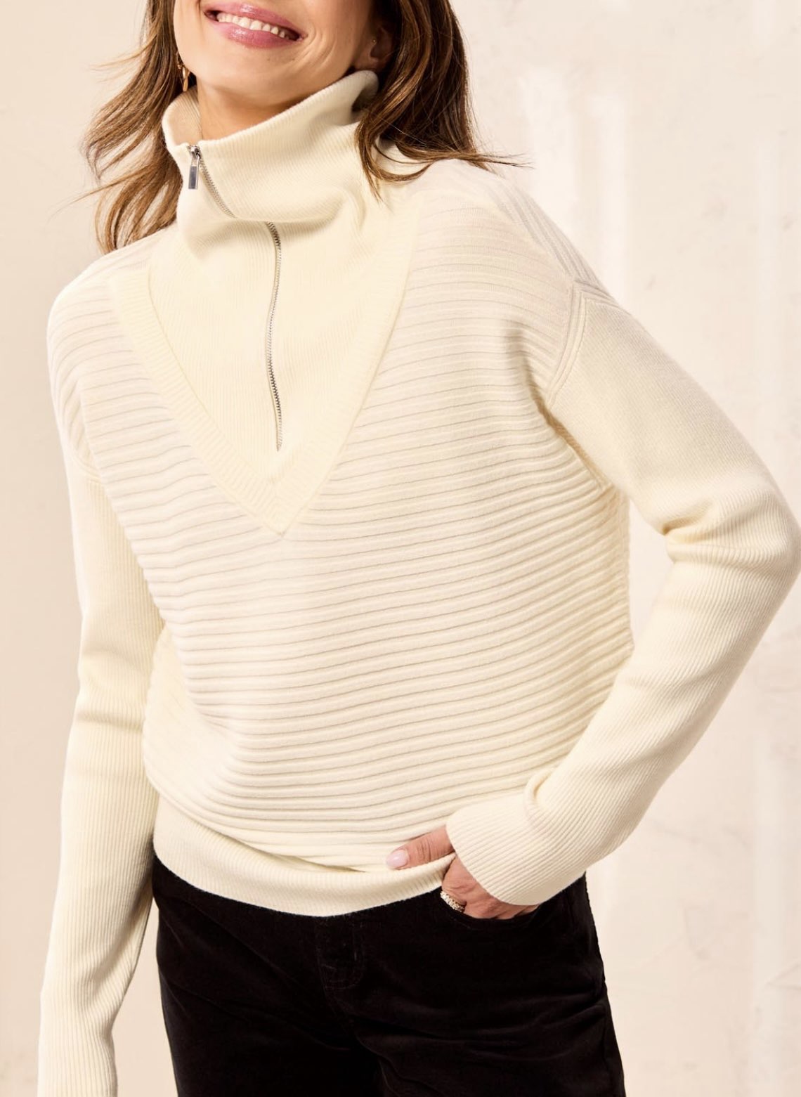 A woman wearing a cream ribbed knit mock neck sweater with a short front zipper, paired with black pants, against a light beige background.