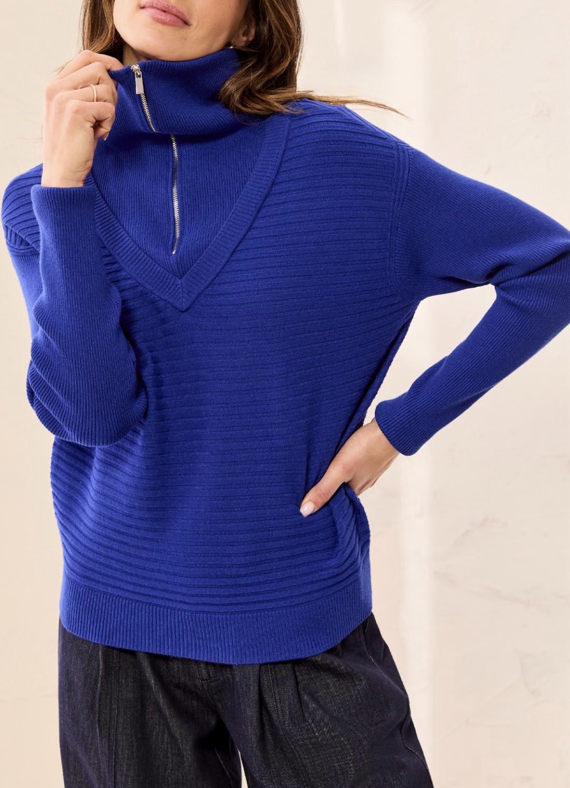 A woman wearing a royal blue ribbed knit sweater with a high mock neck and a silver zipper detail at the collar, paired with dark pants.