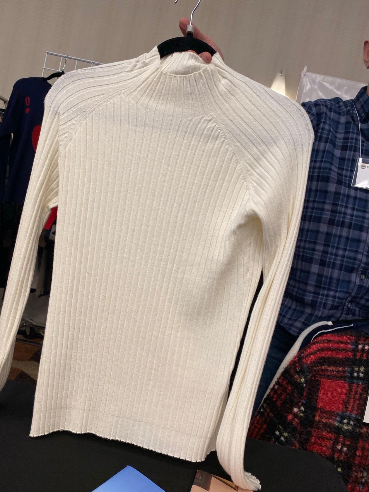An off-white ribbed knit sweater with a mock neck and long sleeves, held on a black hanger against a background of other clothing.