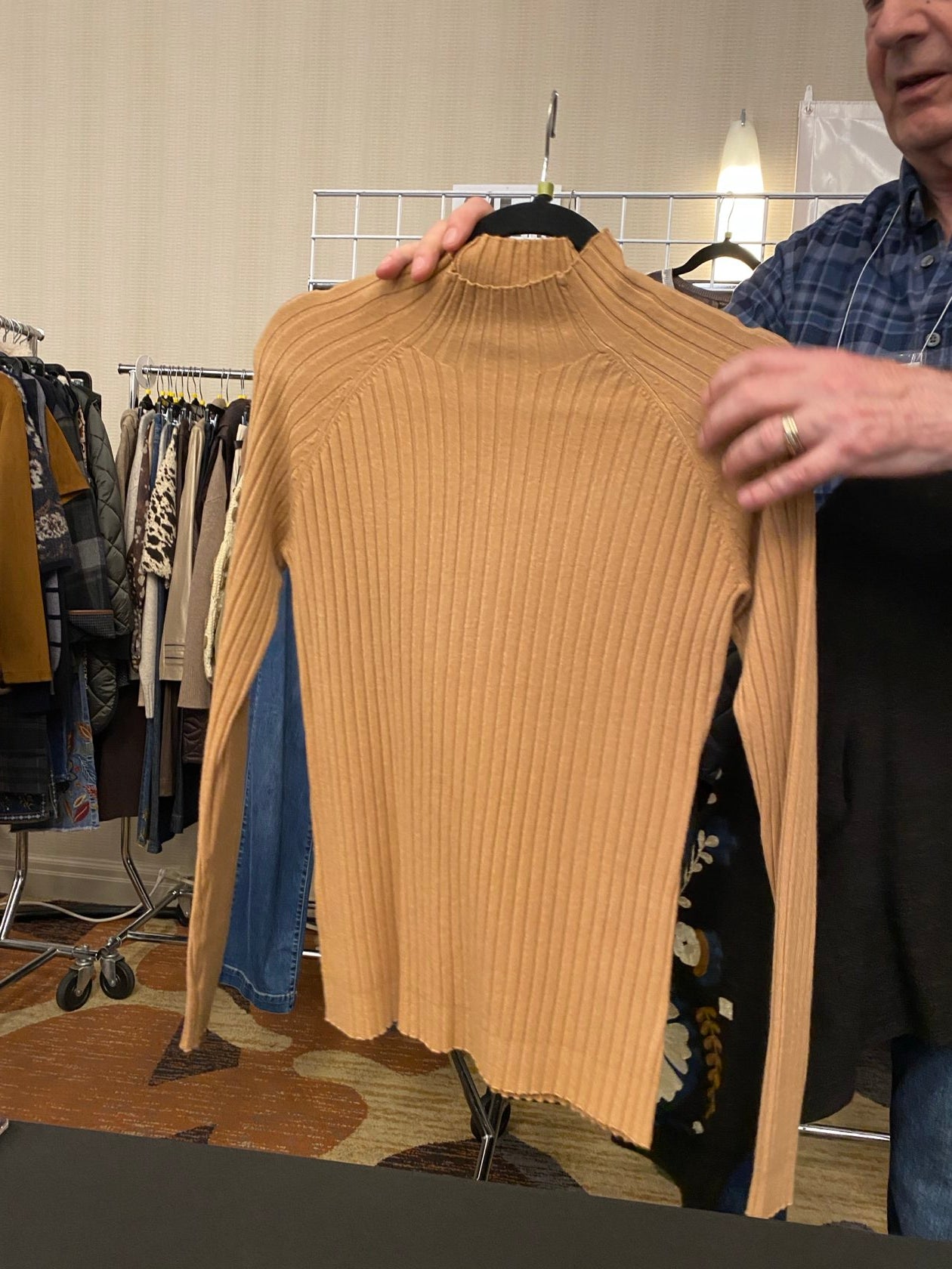 A person holding up a ribbed knit sweater in a warm camel tan color, showing the front of the garment. The background shows a clothing store interior with racks of clothes.