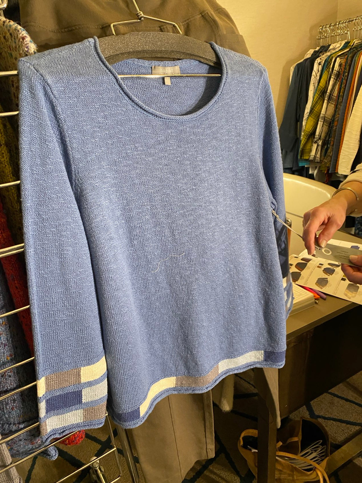 A light blue knit sweater with a striped border in cream, navy, and beige, laid flat on a clothing rack with a person's hands visible in the background.