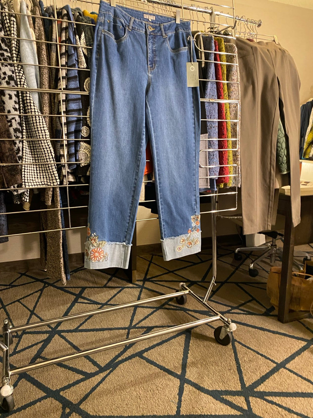 A pair of folded blue jeans with floral cuff details is displayed on a metal clothing rack alongside other folded garments in an indoor setting with a patterned carpet.