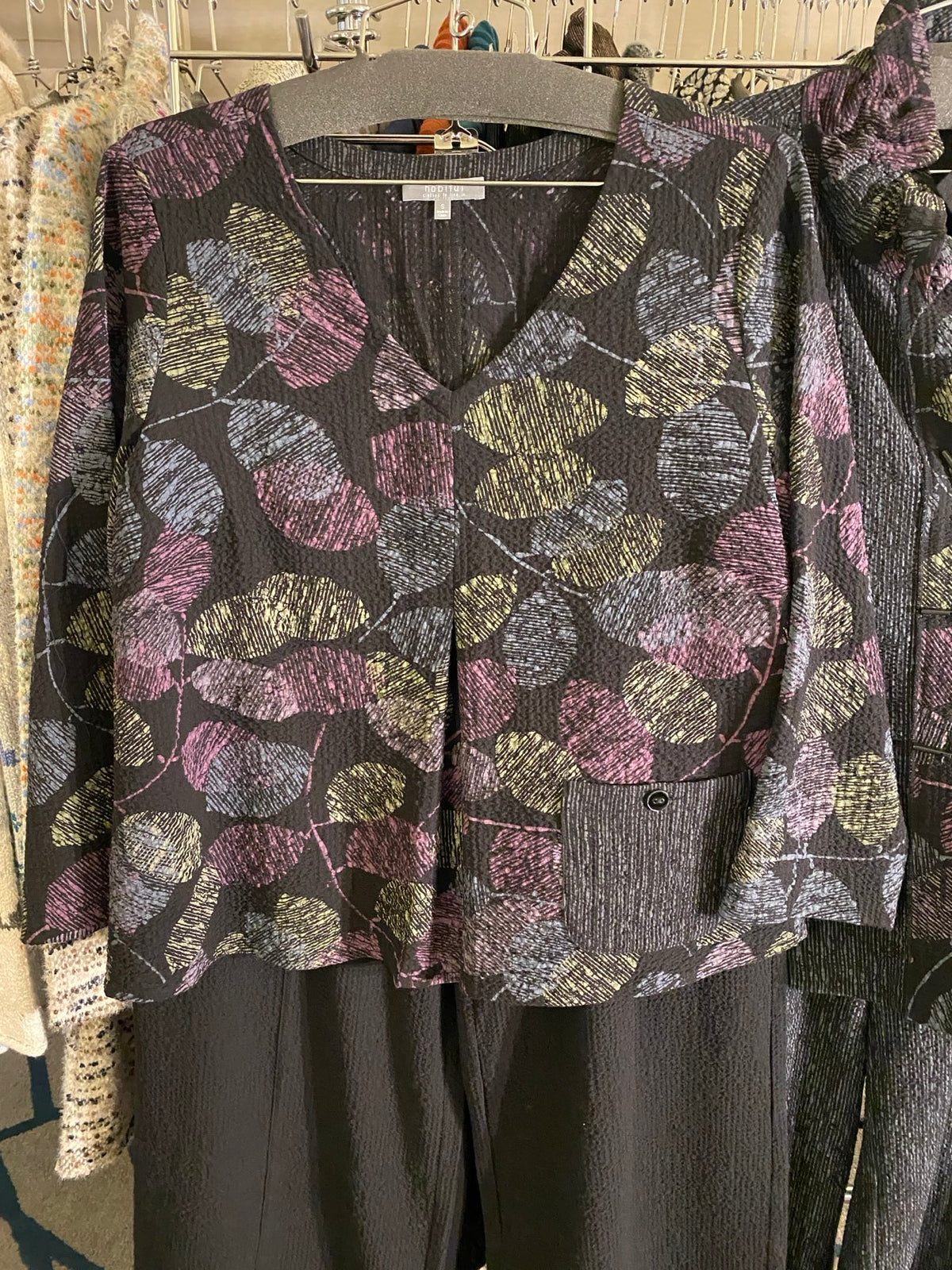 A folded dark gray or black top with a textured leaf pattern in dusty pink, olive green, and beige, displayed on a hanger next to a dark solid garment.