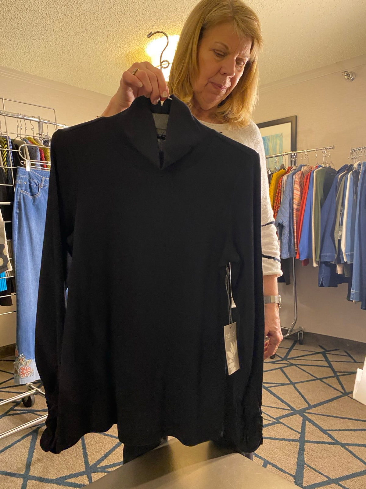 A woman holding up a black long-sleeve top or sweater in a retail store, with clothing racks and a patterned carpet visible in the background.