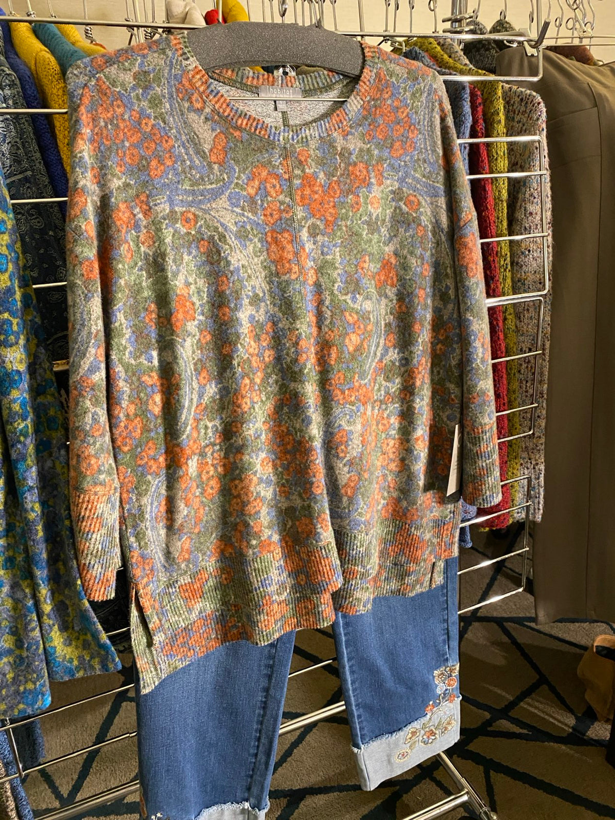 A sleeveless knit top with a colorful paisley and floral pattern in orange, blue, green, and beige, layered over a pair of blue jeans, displayed on a metal rack.