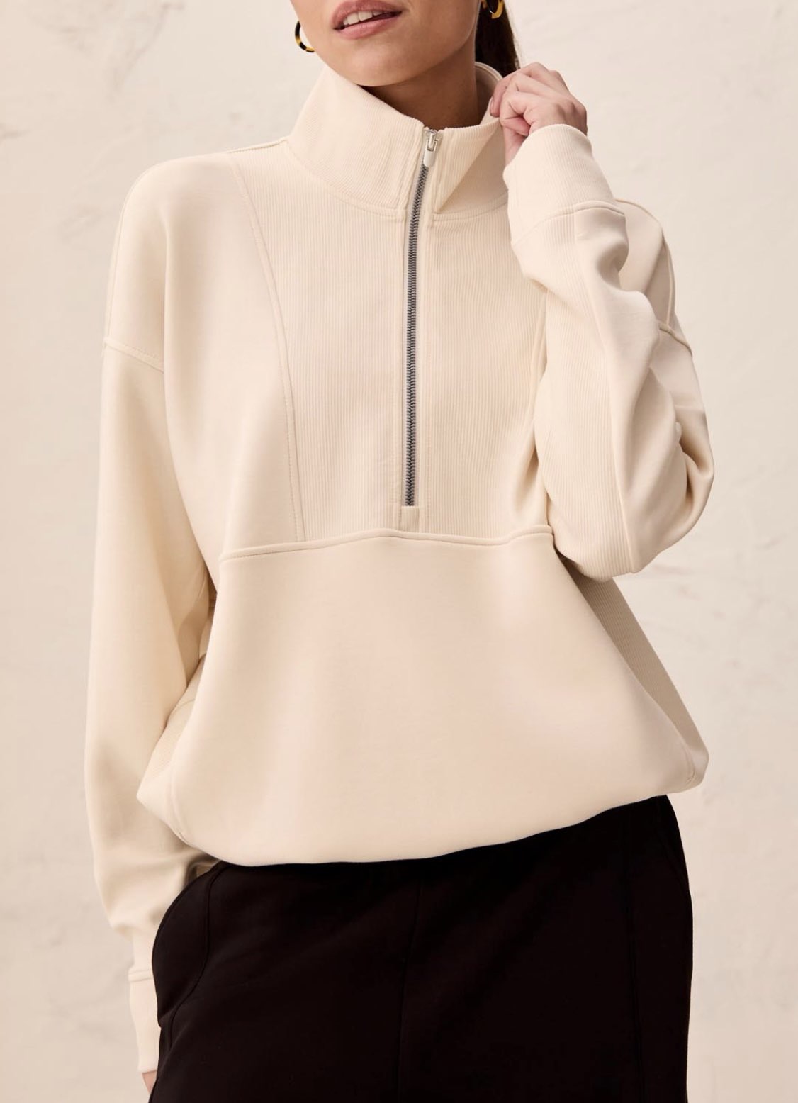 A woman wearing a cream-colored quarter-zip pullover sweatshirt with structured shoulders and black pants, standing against a light textured wall.