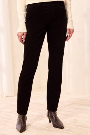 A person wearing slim black ankle-length pants with a cream ribbed top and dark pointed-toe ankle boots, standing against a light cream curtain and pale wood floor.