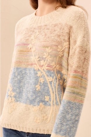 Close-up of a person wearing a cream sweater with floral patterns and color-blocked sleeves.