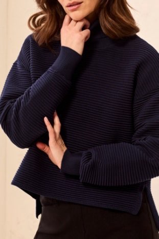 A woman wearing a dark navy ribbed knit mock neck sweater with long sleeves, paired with black trousers, shown in a close-up shot against a neutral beige background.