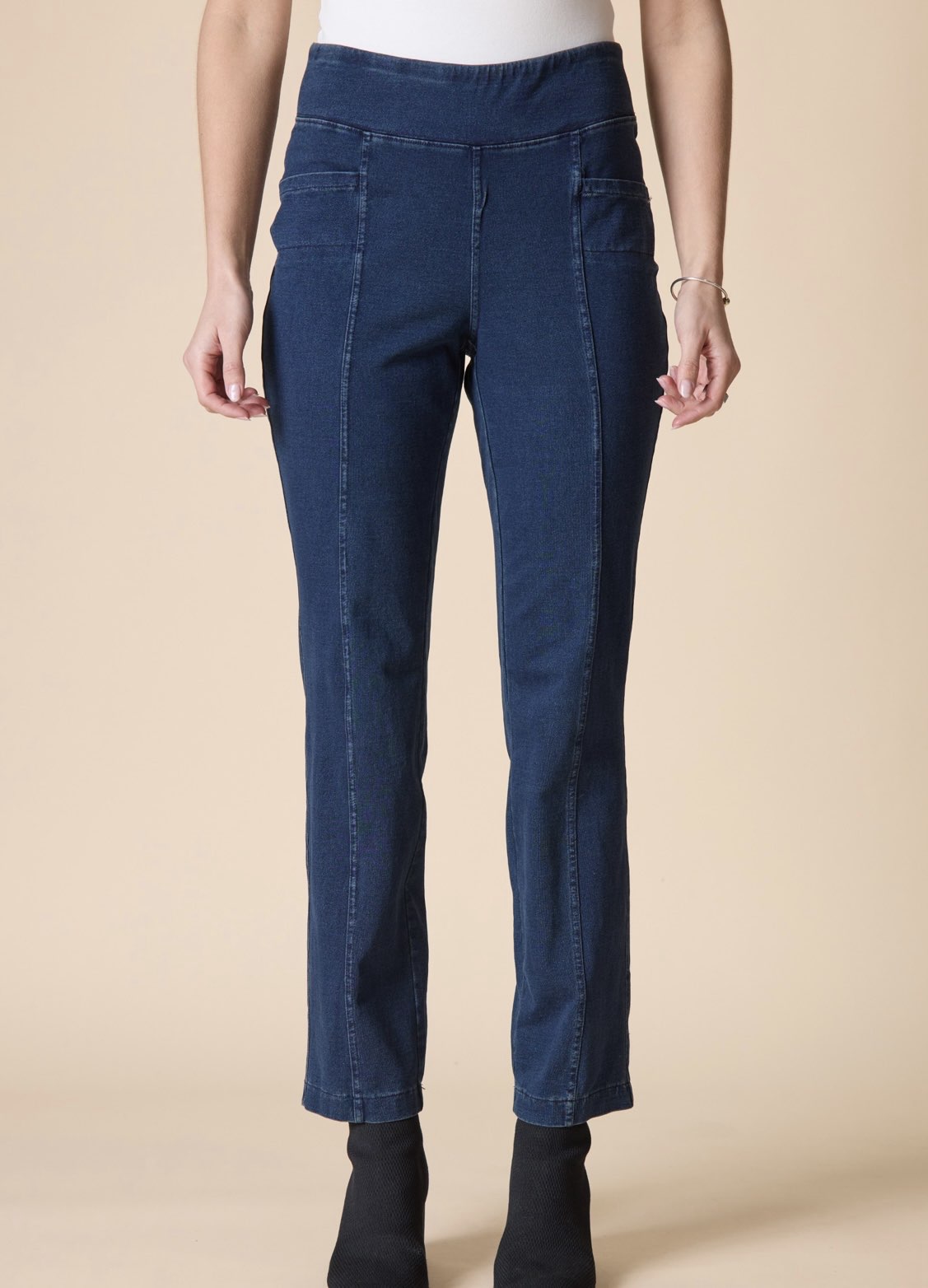 A model wearing dark blue denim jeans with a wide waistband, front patch pockets, and a straight-leg fit, paired with black ankle boots against a solid beige background.