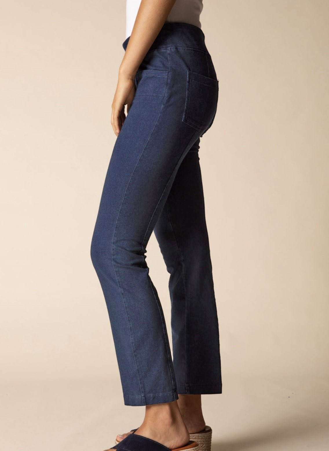 A side profile view of a person wearing dark blue cropped flare jeans, paired with a white top and brown espadrille sandals, against a solid beige studio background.