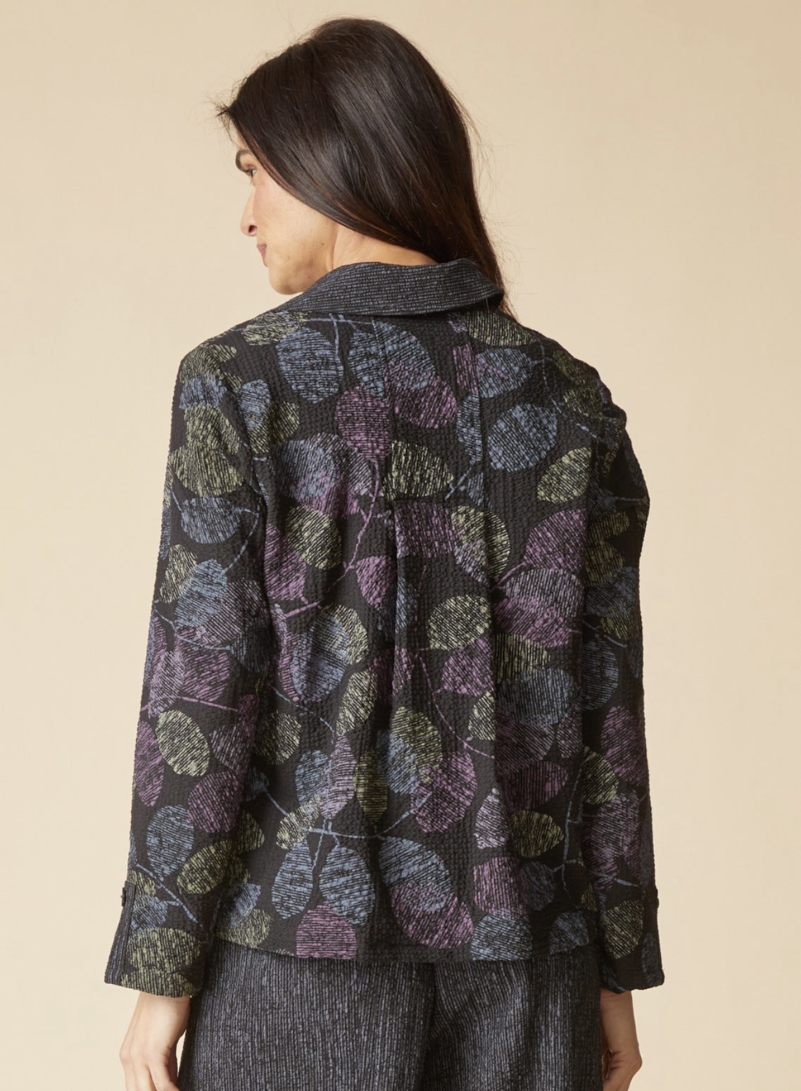 A woman shown from the back wearing a dark charcoal gray jacket with a textured leaf pattern in purple, olive green, and blue, paired with dark gray trousers, against a solid beige background.
