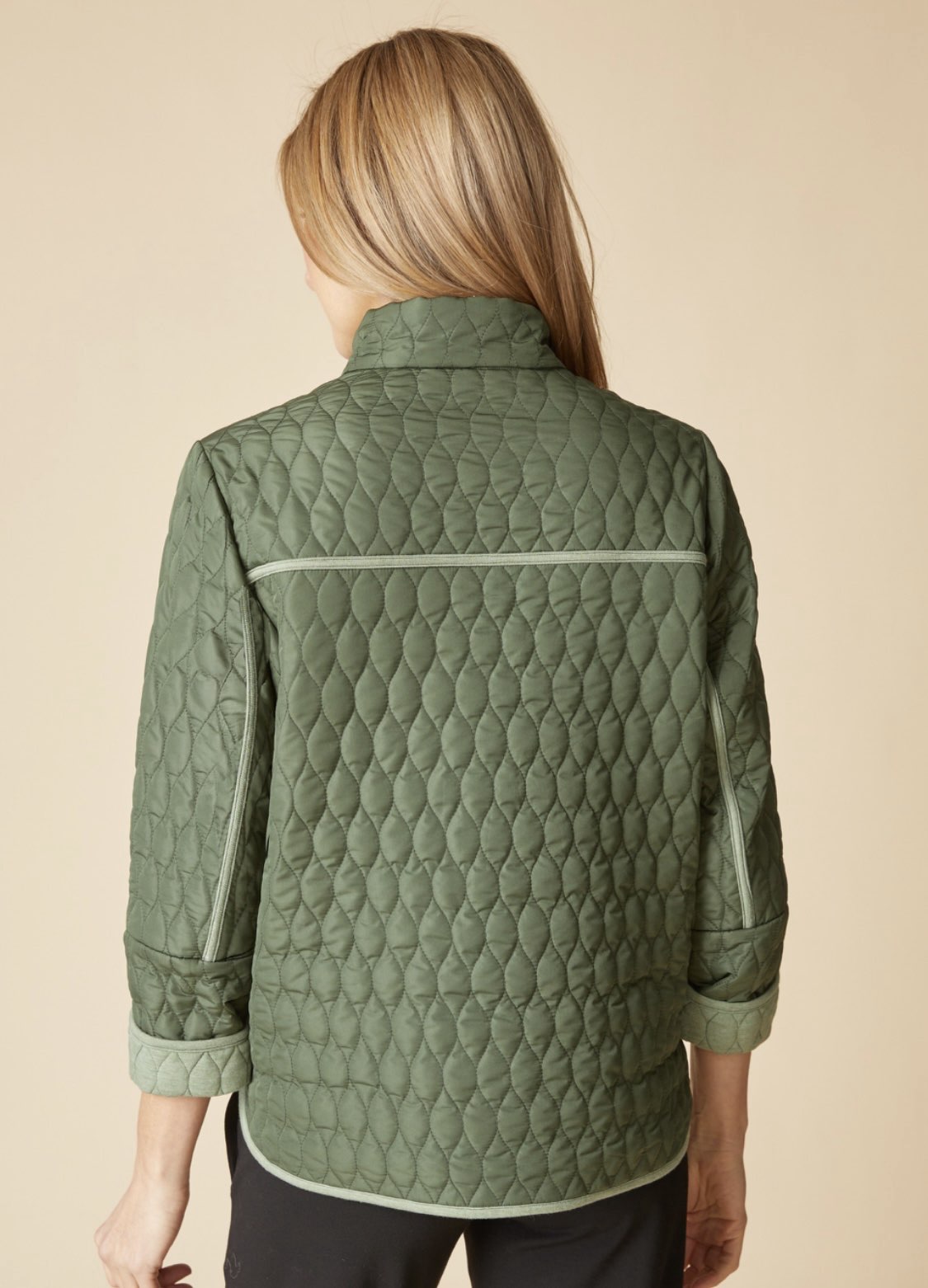 A woman viewed from the back wearing a quilted sage green jacket with a high collar and subtle contrast piping, set against a solid beige background.
