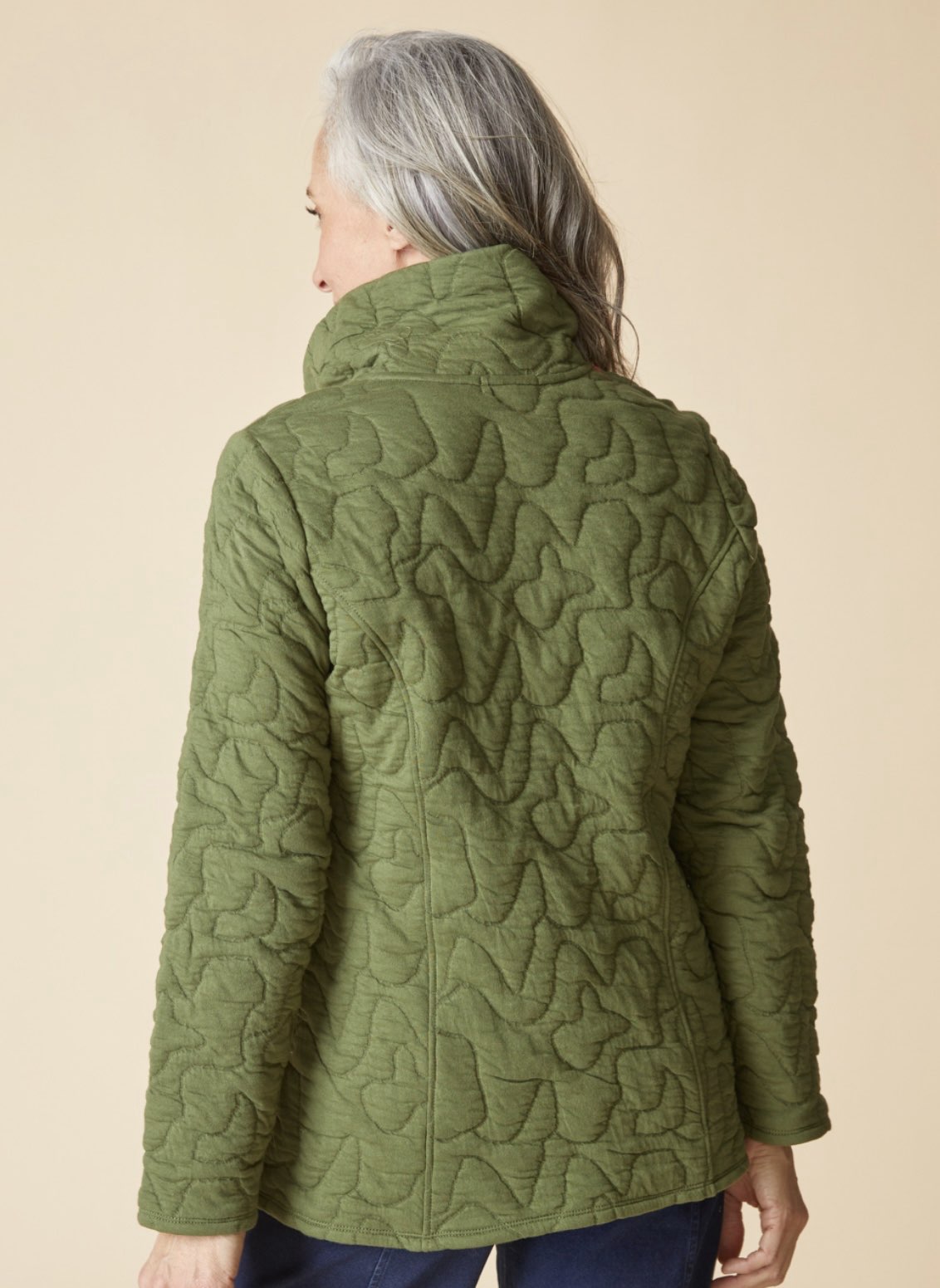 A woman with gray hair is shown from the back wearing an olive green quilted jacket with a high stand-up collar, set against a solid beige background.