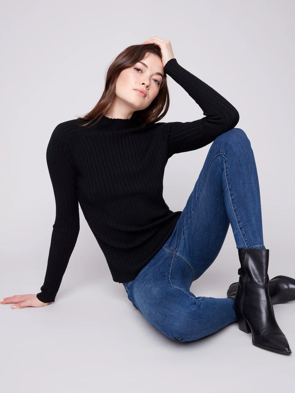 A woman wearing a black ribbed long-sleeve mock neck sweater, blue jeans, and black ankle boots, posing on a light gray studio background.