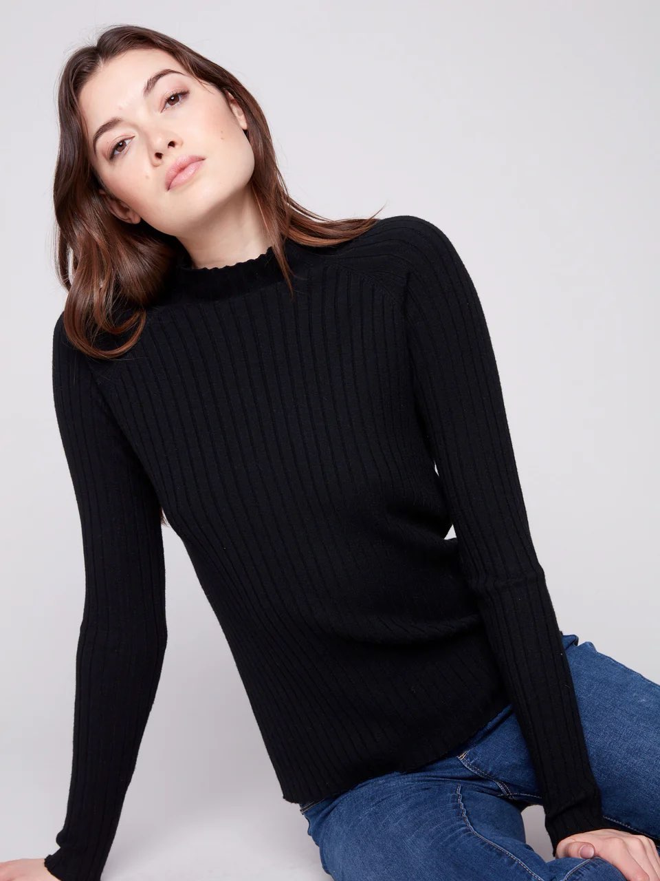 A woman wearing a black ribbed knit mock neck sweater and blue jeans, posing against a light gray studio background.