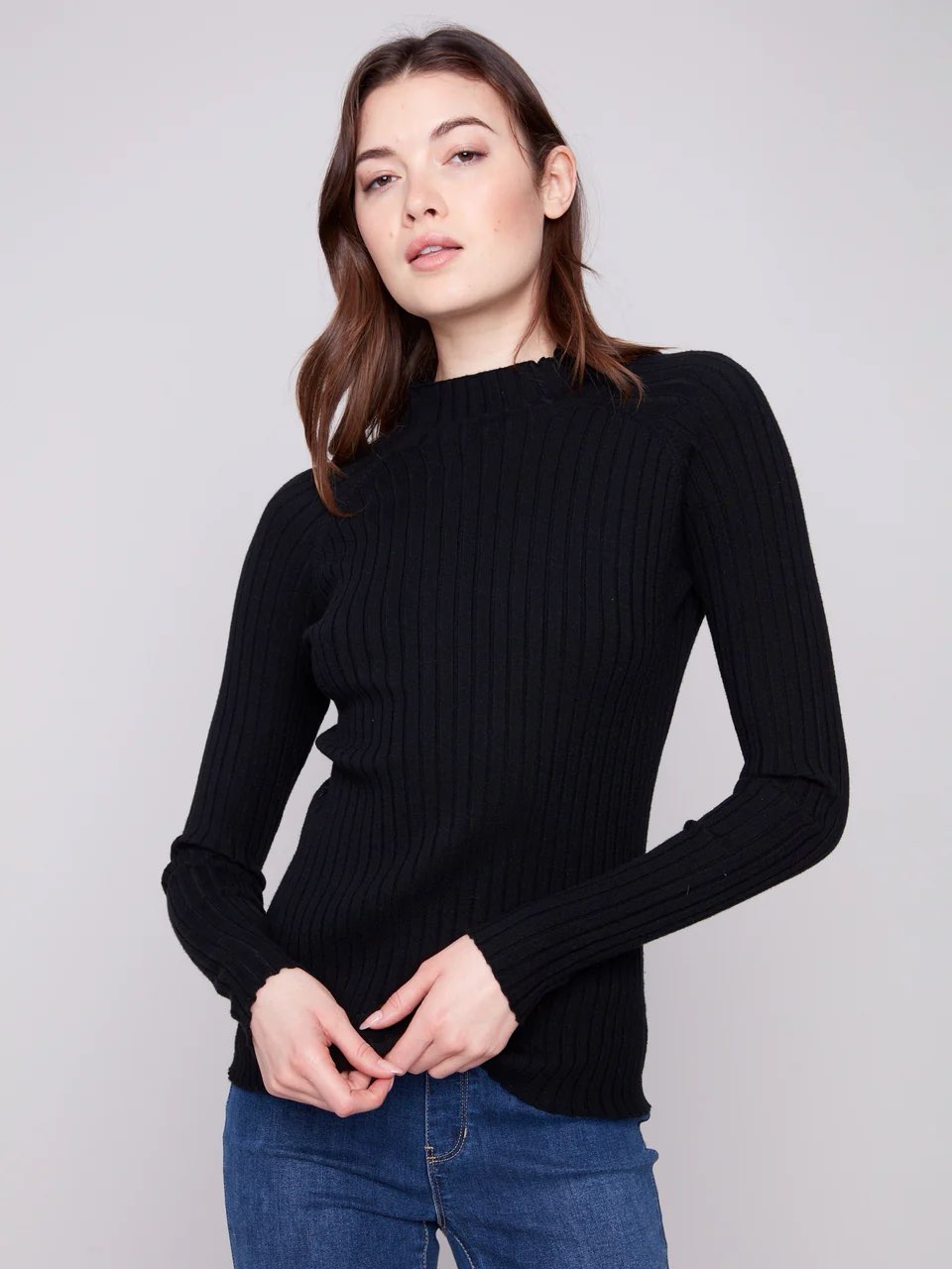 A woman wearing a black ribbed long-sleeve mock neck sweater and blue jeans, standing against a solid light gray studio background.