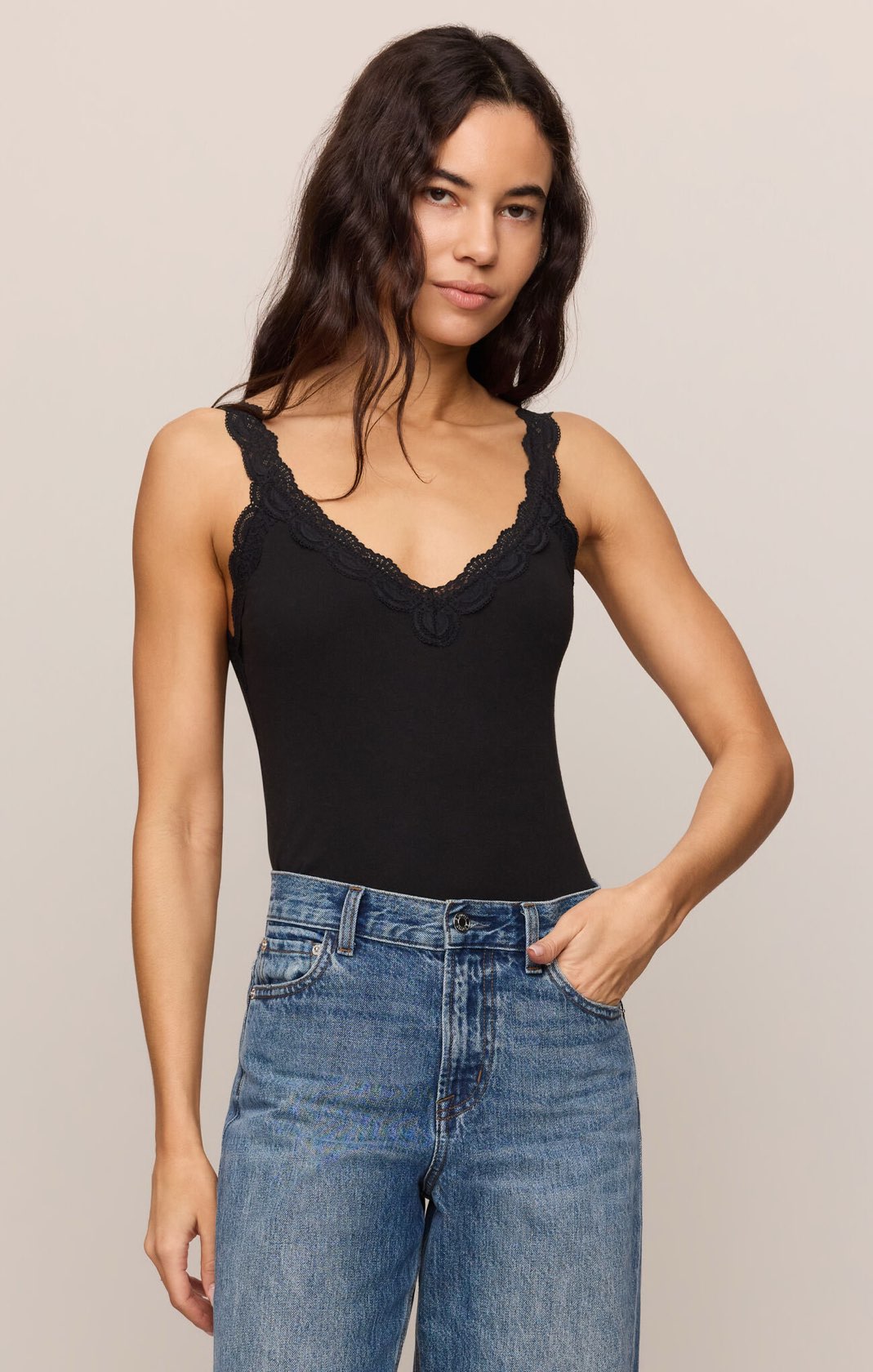 Woman wearing a black tank top with lace details and blue jeans on a beige background
