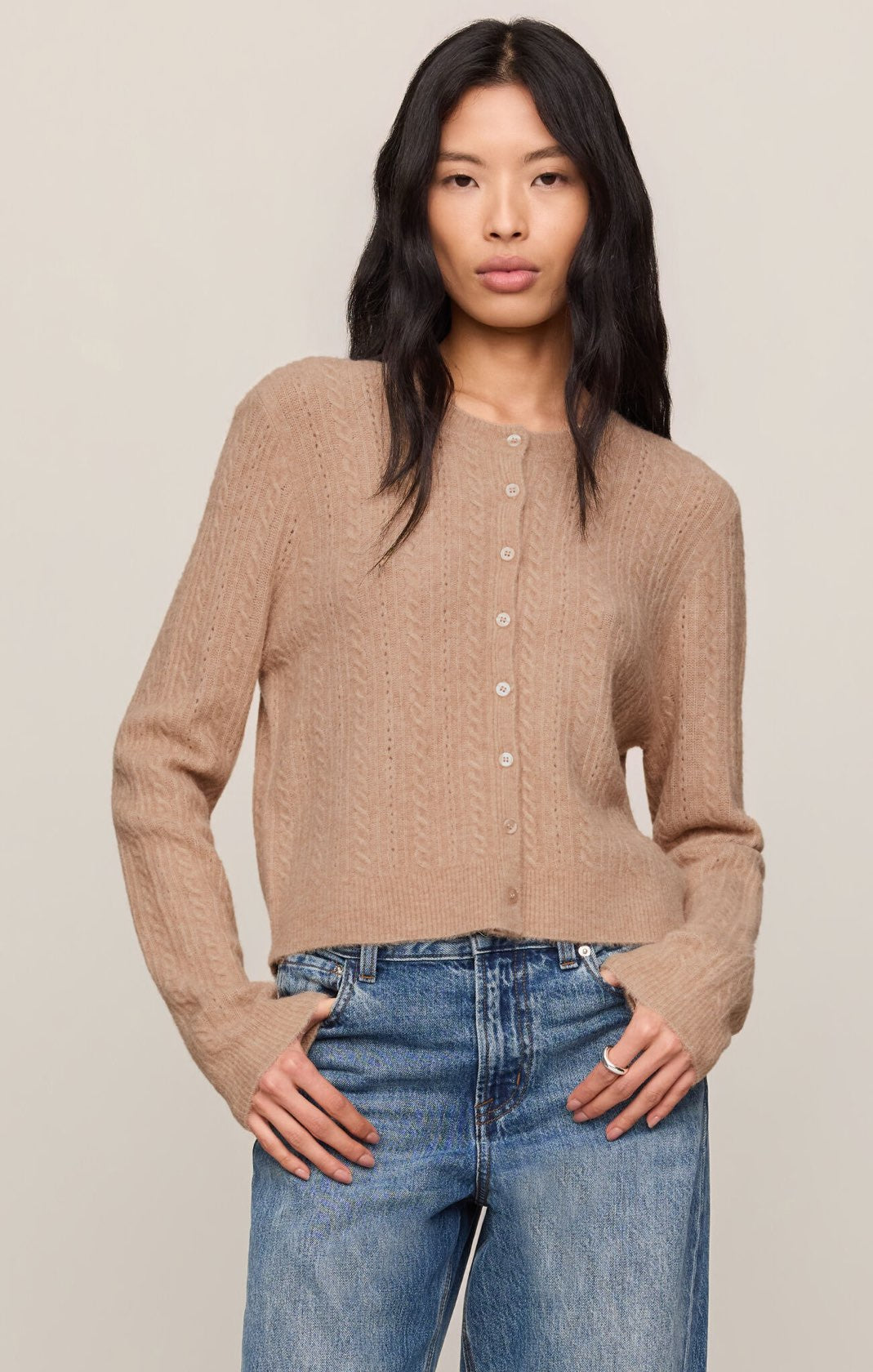 A woman modeling a cropped, long-sleeve knit cardigan in a warm taupe-beige color, paired with blue jeans, against a solid light beige background.