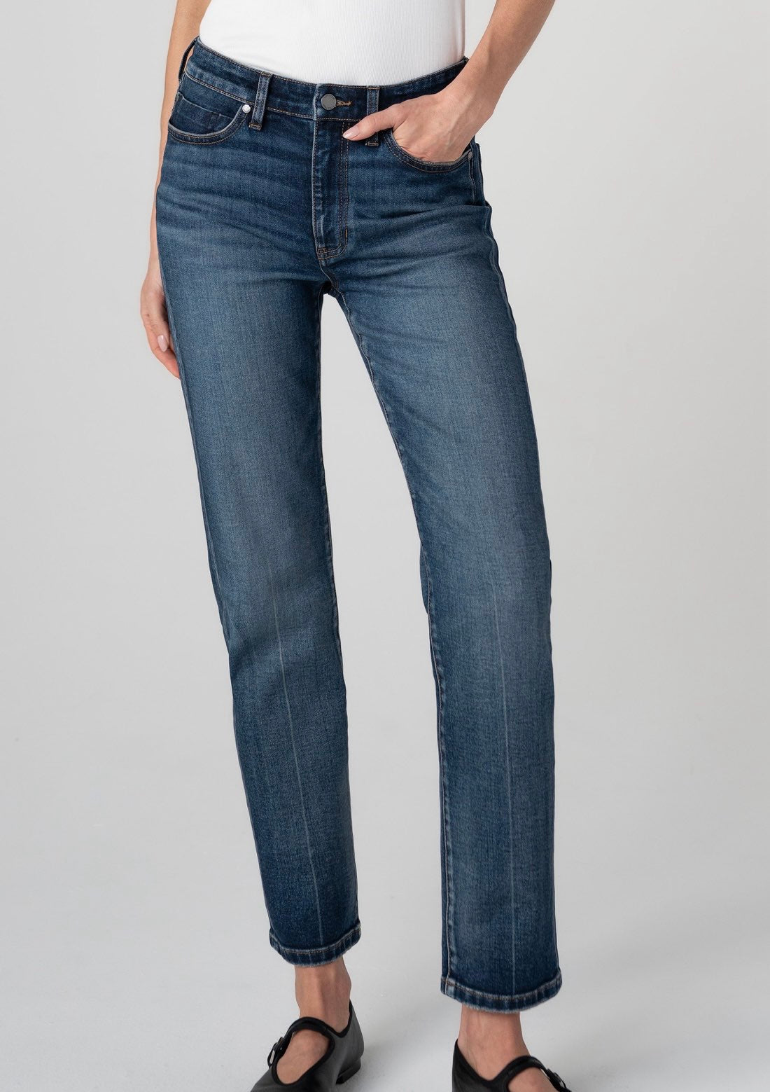 A person wearing dark blue straight-leg denim jeans with a white top and black Mary Jane shoes, shown from the waist down against a light gray studio background.