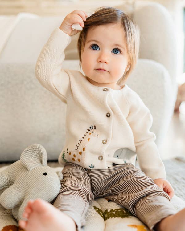 Baby sitting on a soft surface with a plush toy, wearing a white cardigan with animal prints.