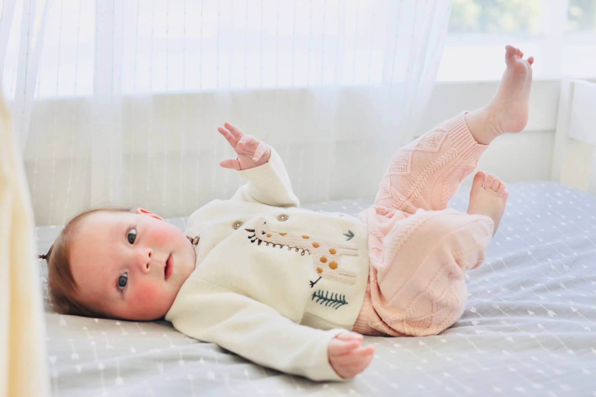 Baby lying on a bed wearing a white sweater with a shark design and pink pants.